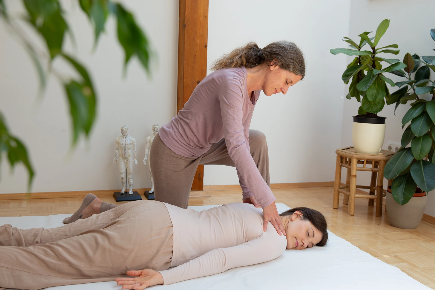 Karin Miedler führt bei einer Frau gerade eine Shiatsu Behandlung durch. Sie übt sanften Druck auf den Rücken aus. Die Frau liegt dabei bekleidet auf einer Matte und wirkt sehr entspannt.