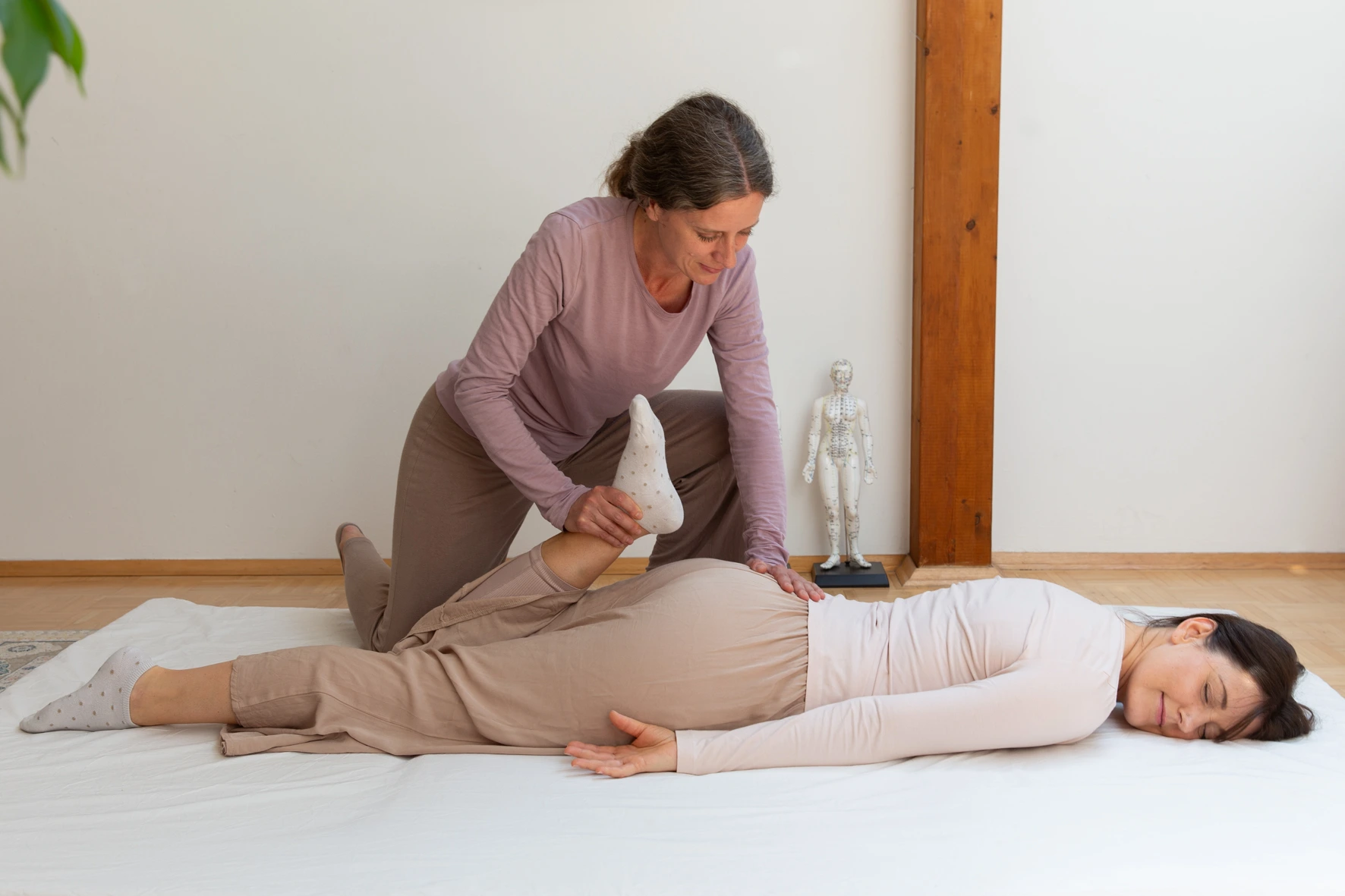 Karin Miedler führt bei einer Frau gerade eine Shiatsu Behandlung durch. Sie dehnt das Bein einer Frau die dabei bekleidet auf einer Matte liegt. Die Frau wirkt sehr entspannt und zufrieden.