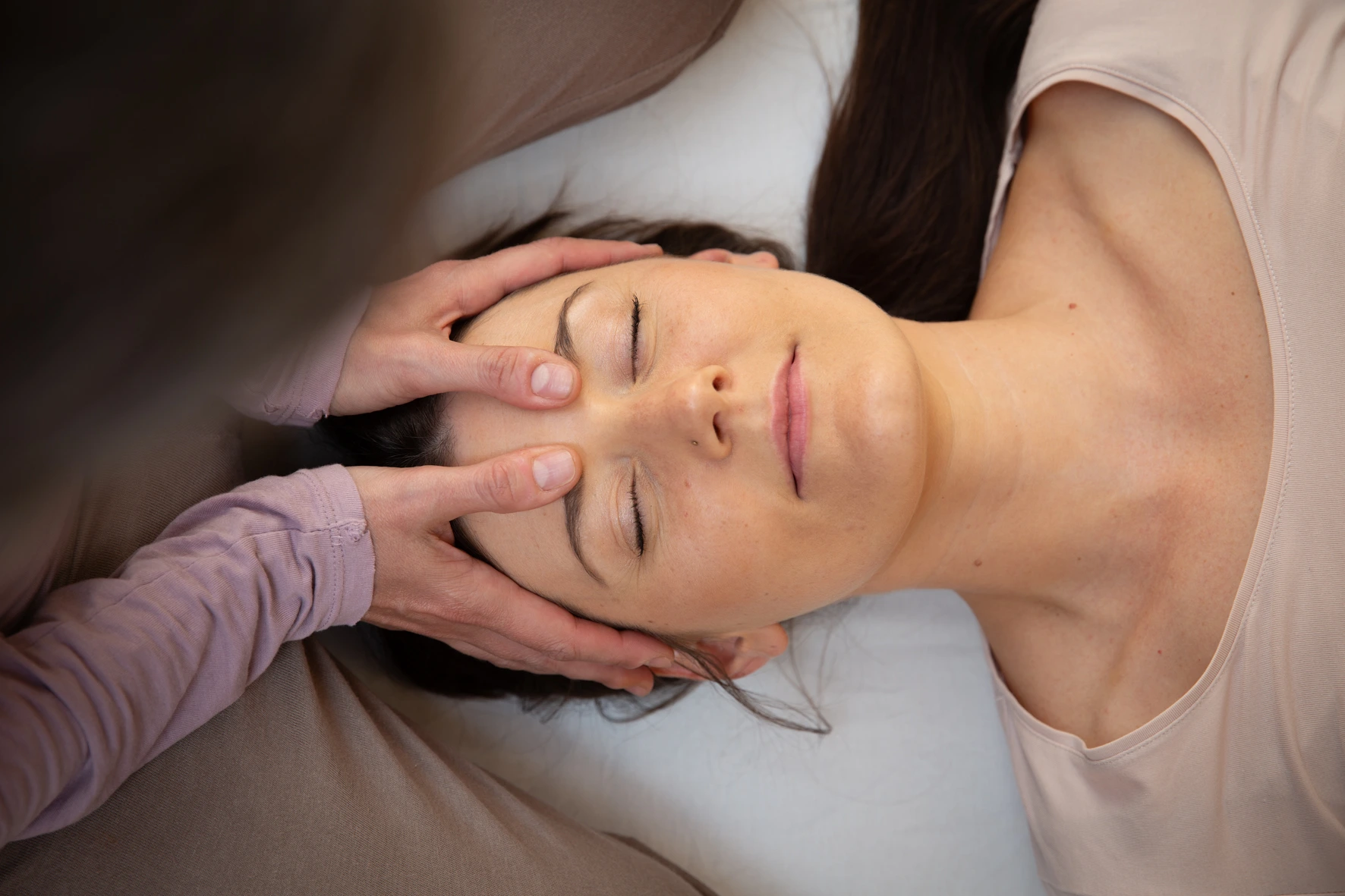 Karin Miedler führt bei einer Frau gerade eine Shiatsu Behandlung durch. Sie übt sanften Druck auf den Ansatz der Augenbrauen aus. Die Frau liegt dabei bekleidet auf einer Matte und wirkt sehr entspannt.